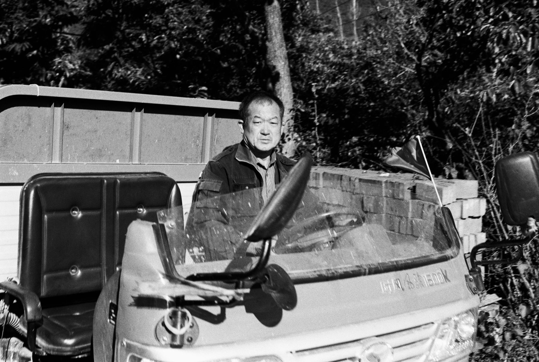 A man leaning on the cab of a small truck beside trees